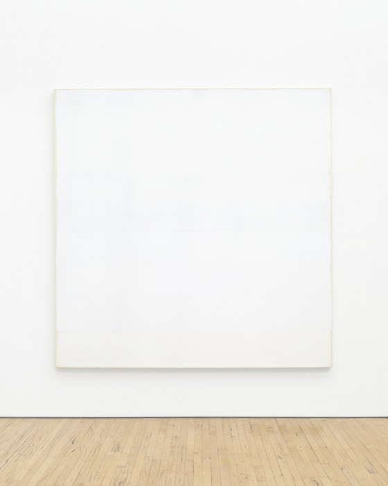 A large, square tan canvas painted white hangs low on a white wall above a wood floor. The canvas is slightly visible around the edges of the paint.