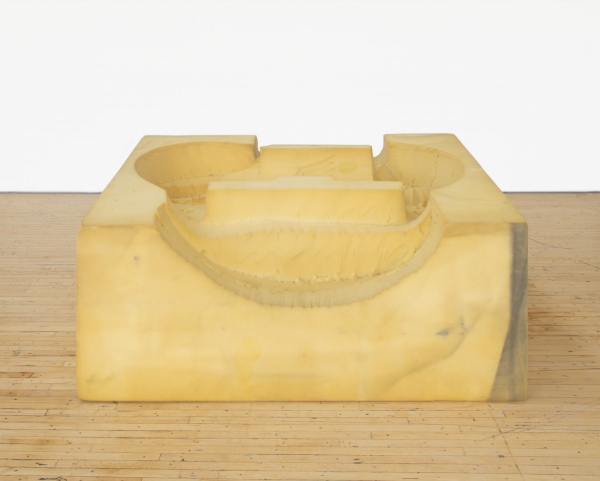 A foam square with curved forms carved into its top rests on a wooden floor in front of a white background.