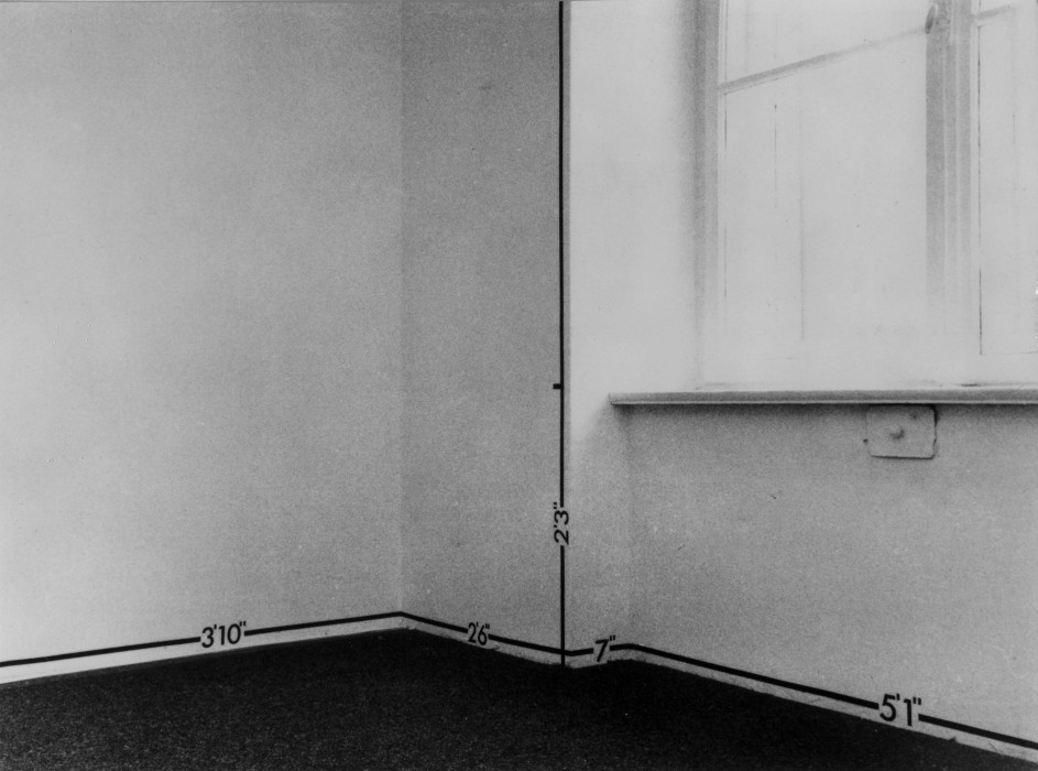 A black-and-white photograph of a white room with a window, the room is marked by lines and notes of measurement.