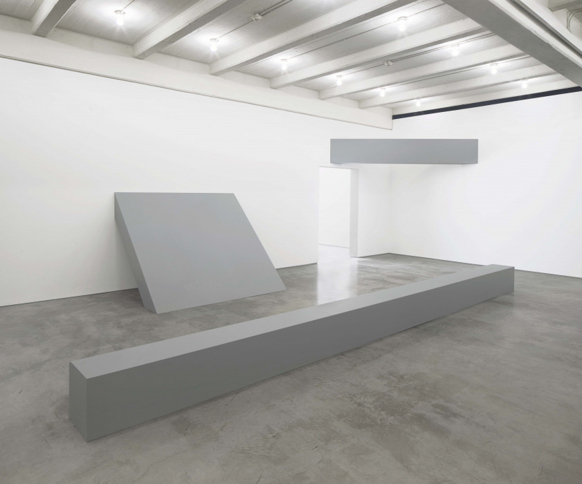 Three large, geometric, grey forms occupy a brightly lit white room. A thin, long rectangular form lays on the concrete floor. A second thin, rectangular form forms a beam between two adjacent walls. The third square-shaped form leans flush against the wall and floor at an incline.