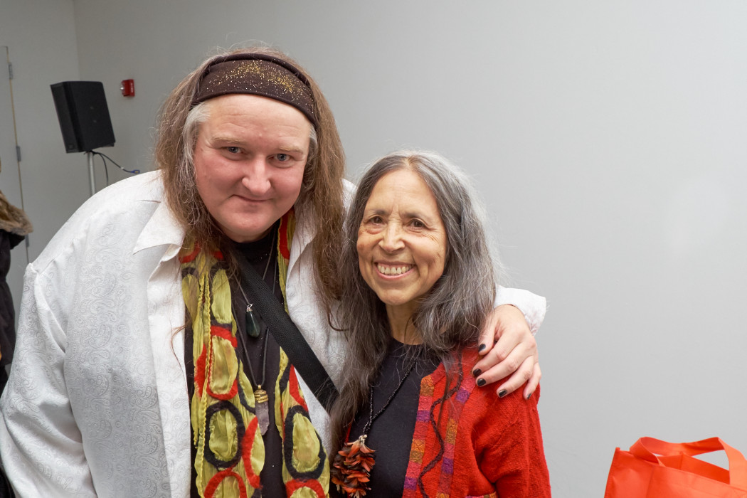 From right to left two poets, one wearing a red cardigan over a black shirt with a pinecone necklace and one wearing a white button up and patterned scarf with their left arm around the shoulders of the other smile a photo against the backdrop of a white-walled room.