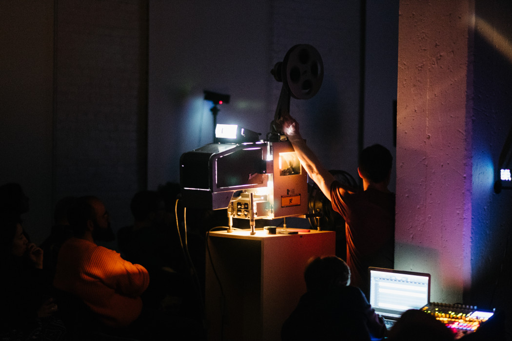 An adult stands operating a film reel projector in a dark room for an audience.