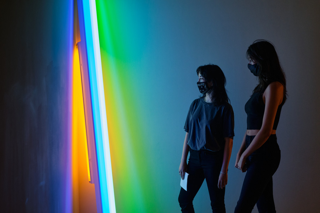 A neon sculpture leans in a corner and glows blue, green, orange, and white. Two people stand near the work and are faintly covered by the light.