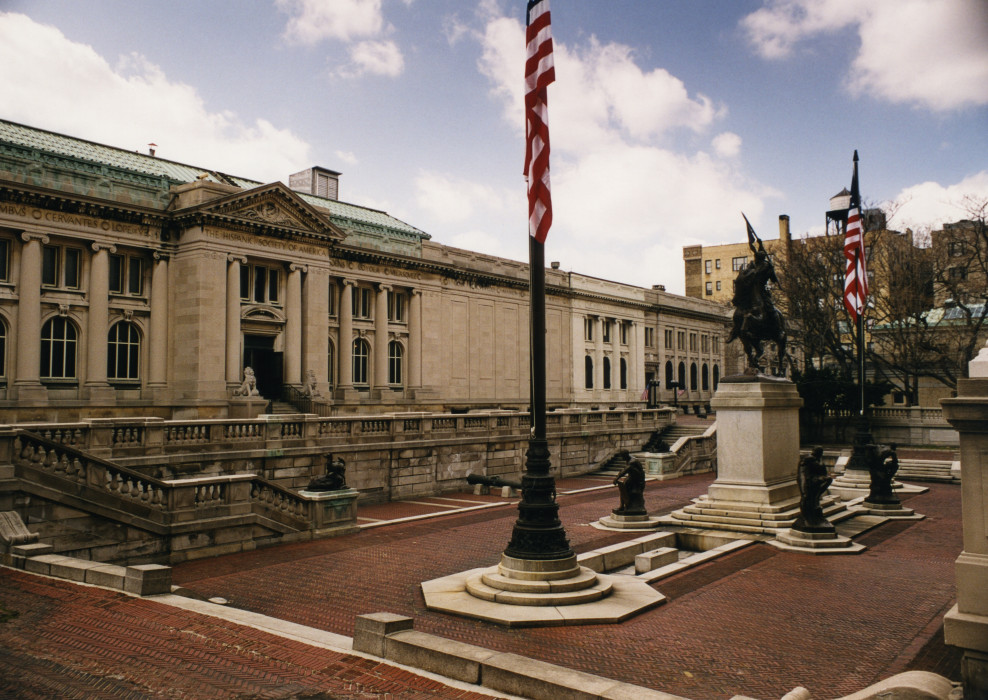A large, long series of buildings behind a terraced, brick-tiled plaza. In the center of the plaza are two American flags atop poles at either end, and a series of statues in between, the largest on a center pedestal with a figure on a horse with their arm raised, holding a flag.