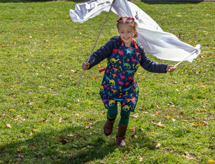 A child runs in a field with large smile on her face while she wears a dinosaur apron. She holds a white flag which flows behind her.