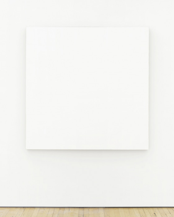 A large, square canvas painted entirely white hangs on a white wall above a wood floor.