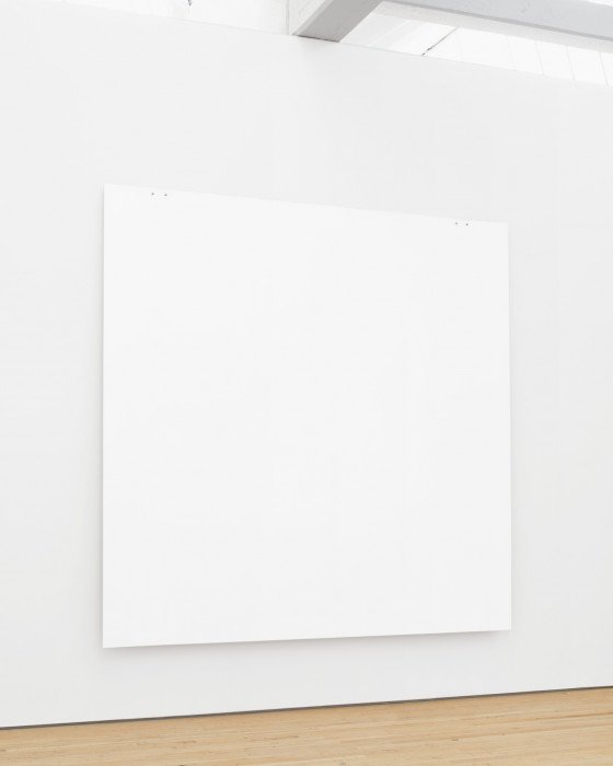 A white square painting hangs low on wall above wood floor using two pairs of tiny bolts on the top edge near the corners. White ceiling beams are visible above the work.