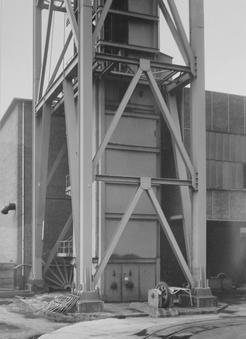 Black-and-white photograph of a large vertical tower encased in an open-air metal support structure, outside a rectilinear brick building.