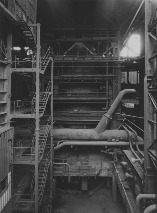 Black-and-white photograph of a multilevel factory interior with platforms, staircases, railings, and a metal support structure surrounding a blast furnace.