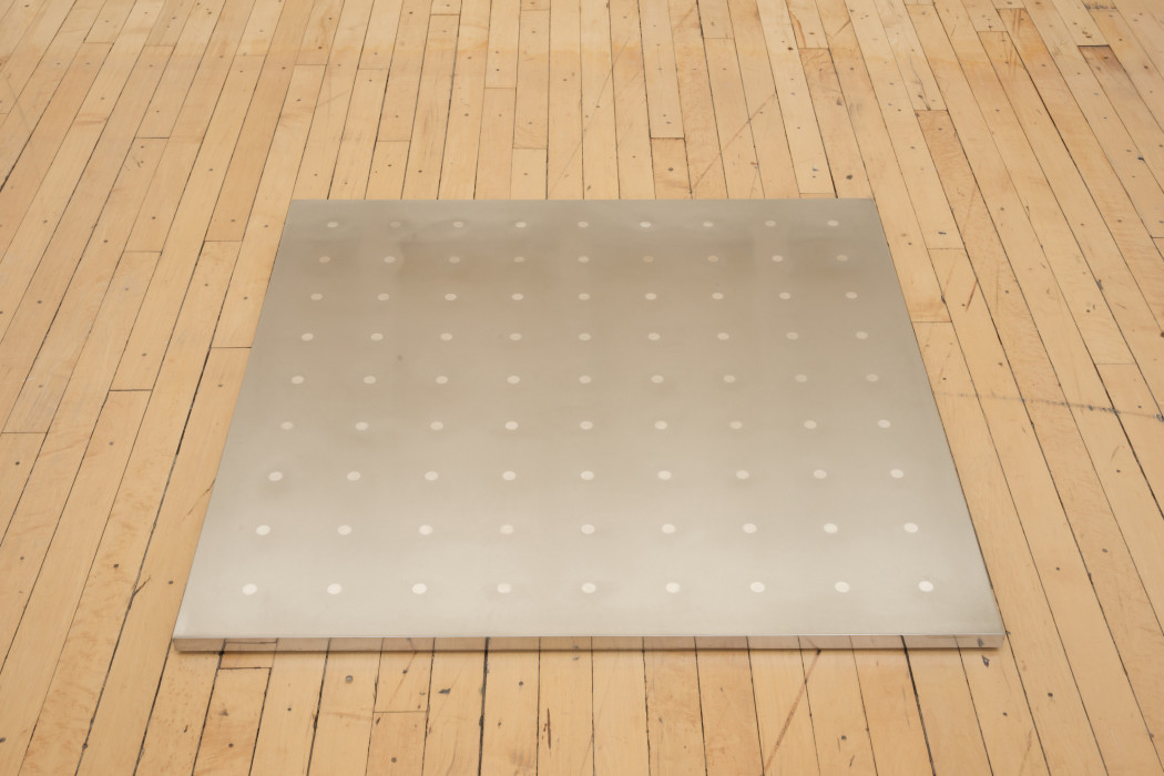 A square silver plaque featuring a grid of silver dots lies flat on a wood floor.