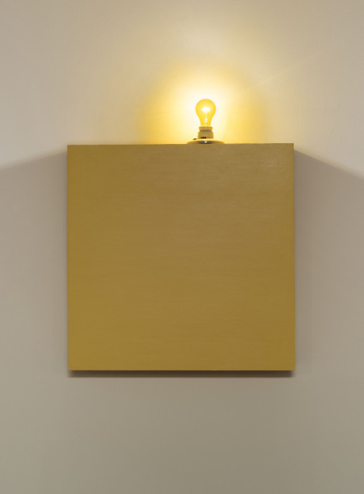 A yellow box topped with a yellow lightbulb hangs on a white wall.