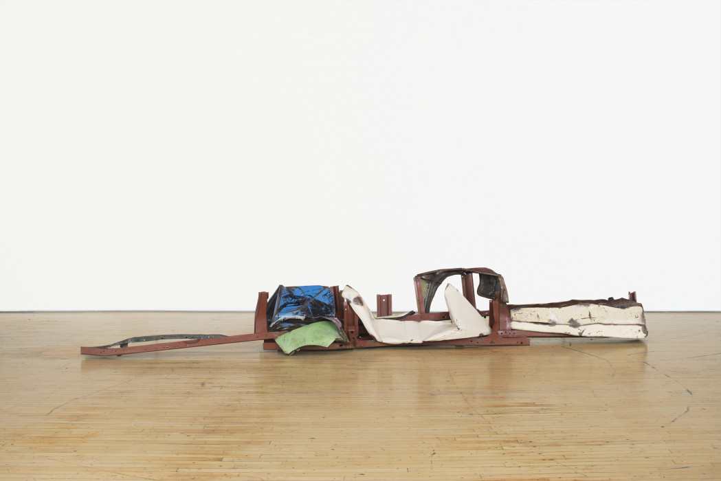 A long, low-lying sculpture made of red, blue, green, and white metal parts rests on a wooden floor.
