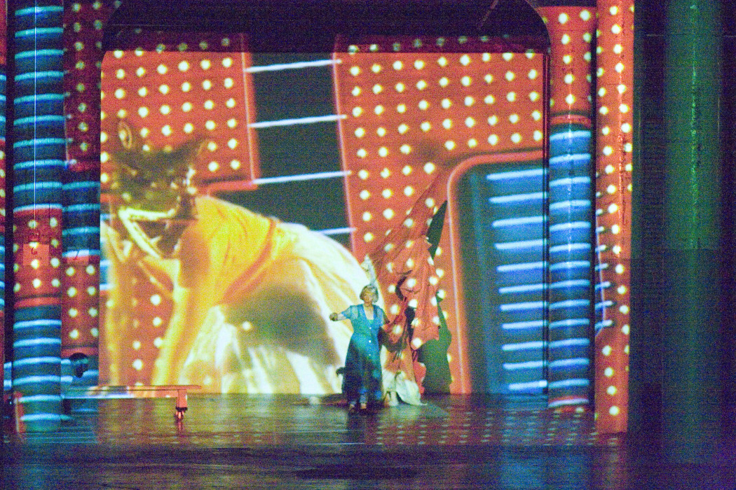 A woman wearing a blue dress stands in front of a screen with projected images of a large cat and red-and-blue shapes.