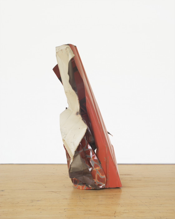 A vertically oriented sculpture made of red, white, and silver-striped red metal parts rests on a wooden floor.