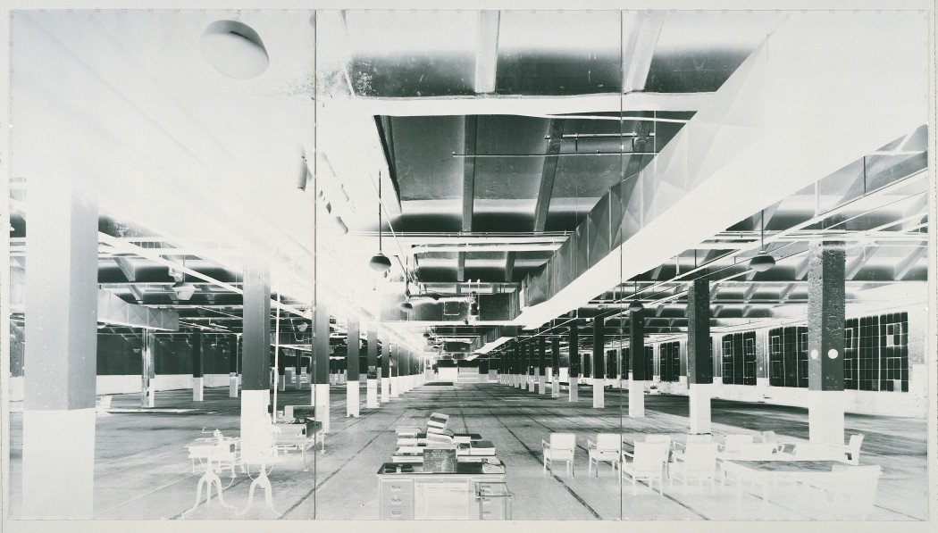 High-contrast, black-and-white photograph of nearly empty, large factory room with chairs, desks, and books in foreground and vertical columns evenly spaced throughout room.