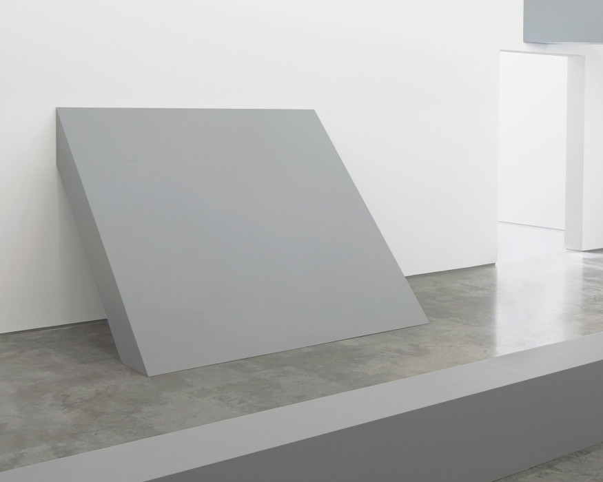 A thick, rectangular, gray sculpture leans against wall.