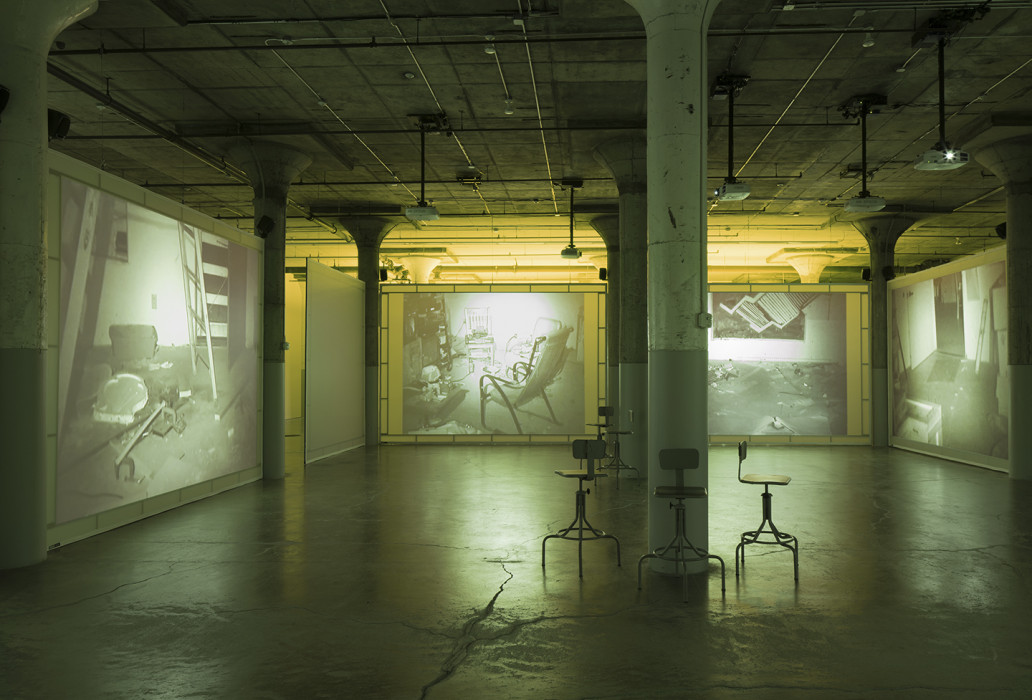 Large industrial space under green light with projection screens serving as walls, displaying images of a small room containing various debris including furniture.