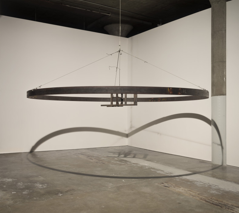 A metal chair in the center of a large circular band, both suspended in an industrial space with concrete floor and white walls onto which the object casts a prominent shadow.