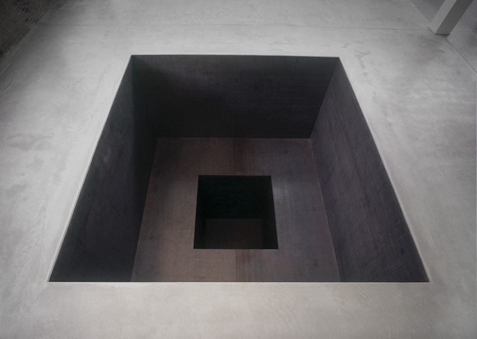 Negative space is carved into a cement floor in the shape of a square. A second smaller square is carved into the first and further recedes into the floor.