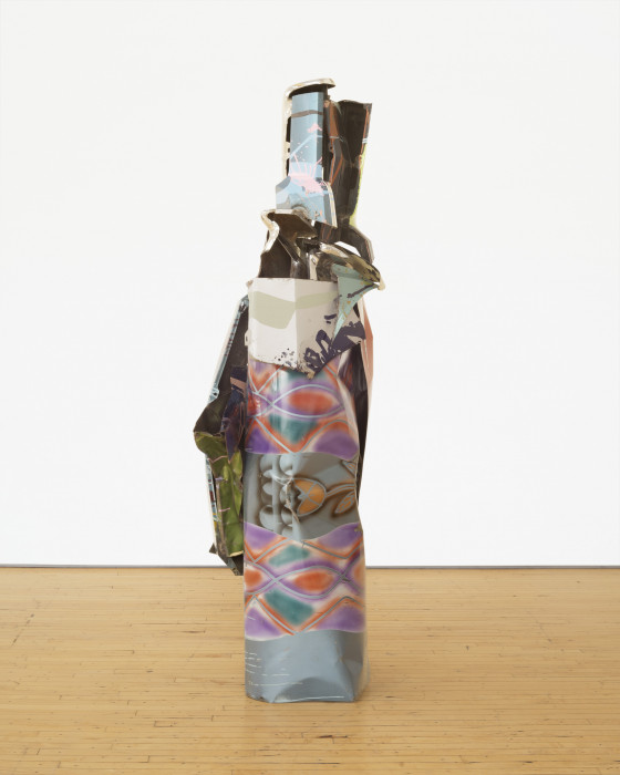 A vertically oriented sculpture made of multicolored metal parts rests on a wooden floor.