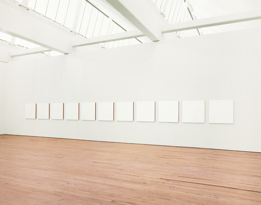Room with eleven evenly spaced, white squares placed along a horizontal white wall above a wooden floor.
