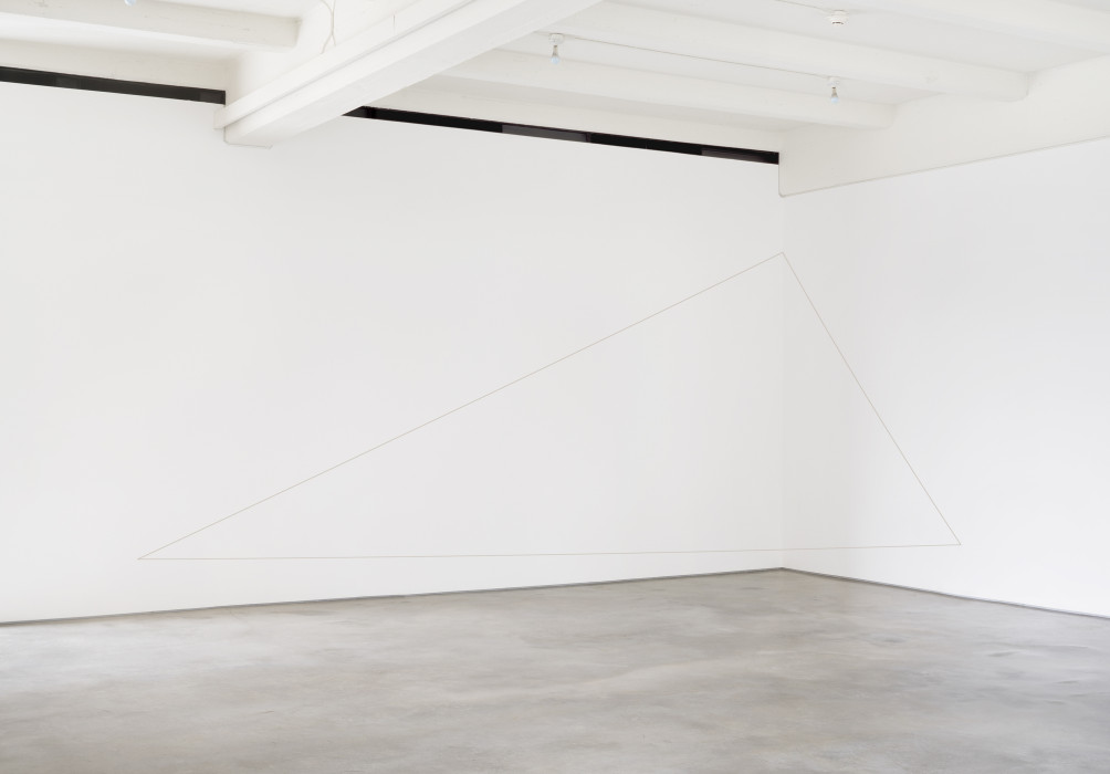 A dark thread suspended from the top of a wall forms a triangle with uneven sides in an industrial gallery space.