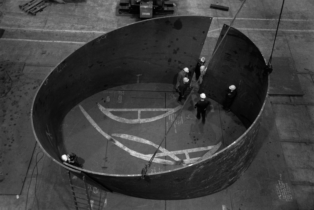 Seven people with safety helmets are gathered around and within two large, curved forms that overlap at one point to create a circular shape in this black-and-white image.