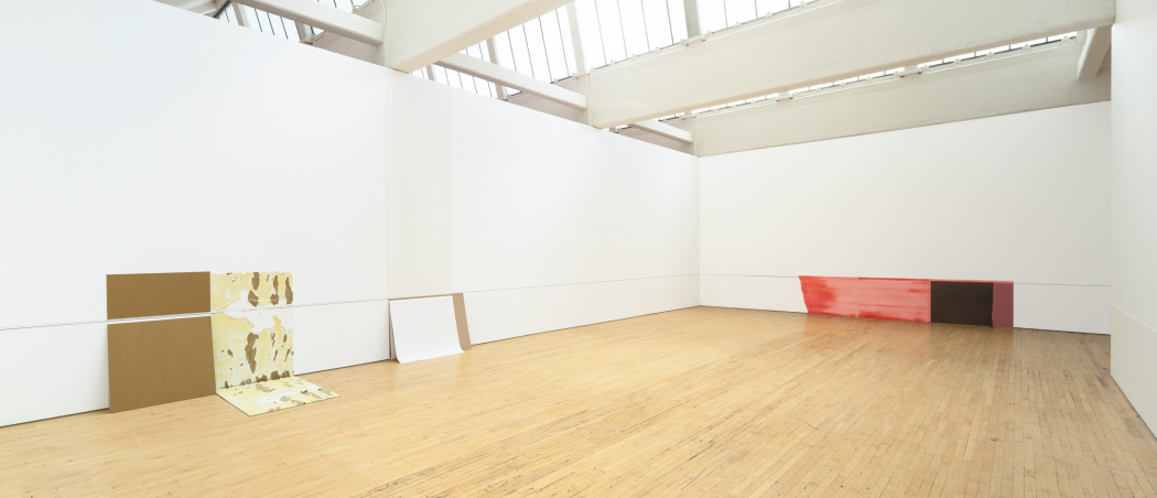 Three rectangular works of various colors, sizes, and materials are placed directly against two connecting white walls that feature a continuous horizontal line a few feet above a wooden floor.