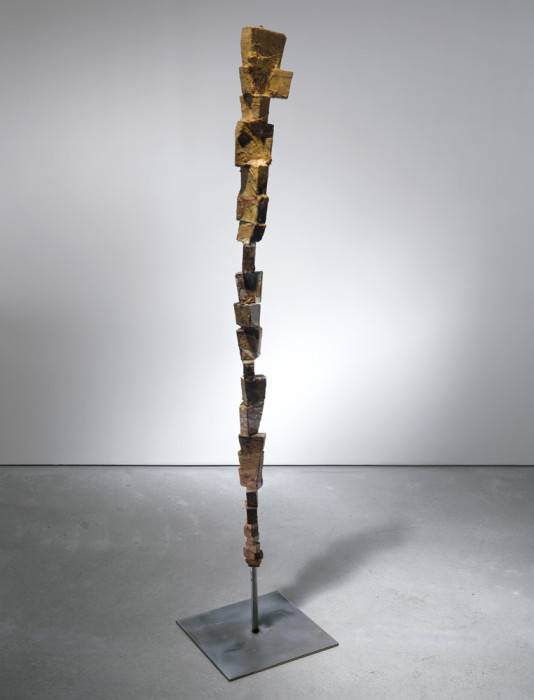 Balanced on a bronze pole and attached to a flat base is a thin vertical sculpture made of several wooden blocks of varying sizes that are stacked on top of one another. Each block is painted in multiple shades of brown.