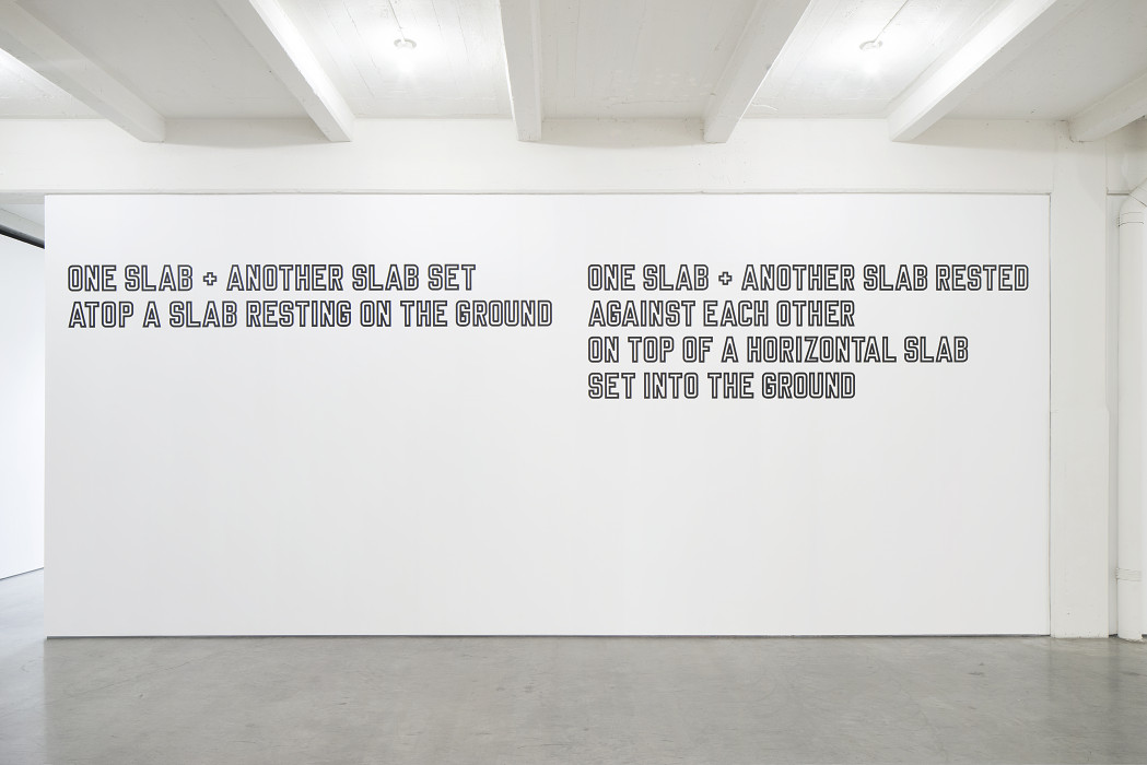 Two paragraphs of white text outlined in black are placed high on a white wall. The text on the left reads: 
