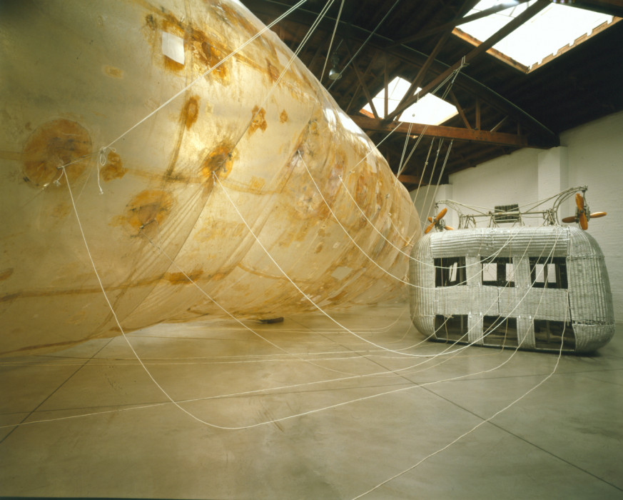 A large, metallic silver, rectangular box with cut-out windows sits to the right of a massive, long horizontal balloon that fills a warehouse space. Atop the silver box sits a bench inside of a metal frame that hangs two orange propeller machines from its ends. White rope connects the box and balloon together at multiple points.