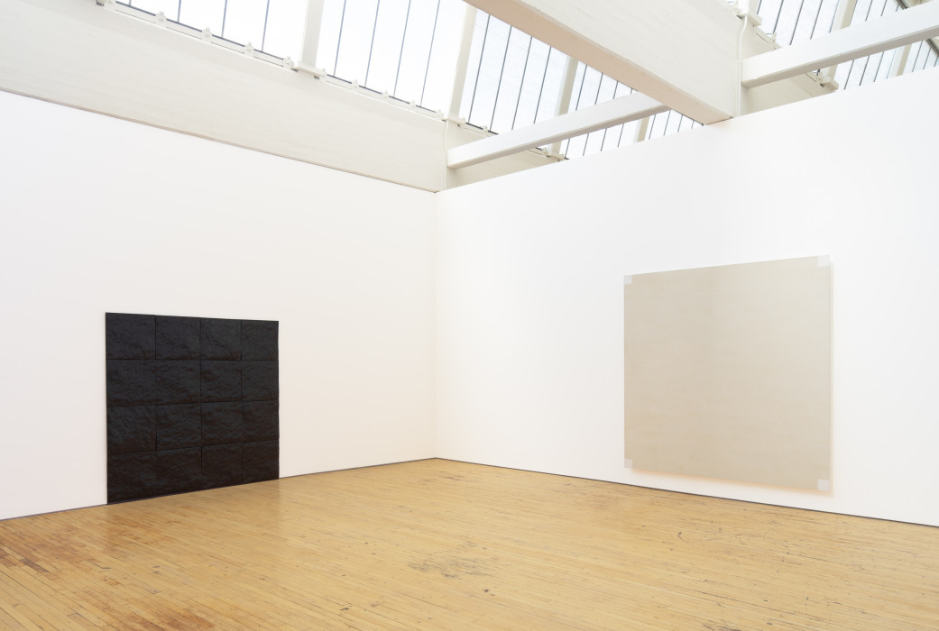 A square four by four grid of shiney dark textuerd tiles hangs on a white gallery wall. The bottom edge touches the gallery's wood floor. On the adjacent wall hangs a larger light grey painting with small white squares painted in the canvas's four corners.