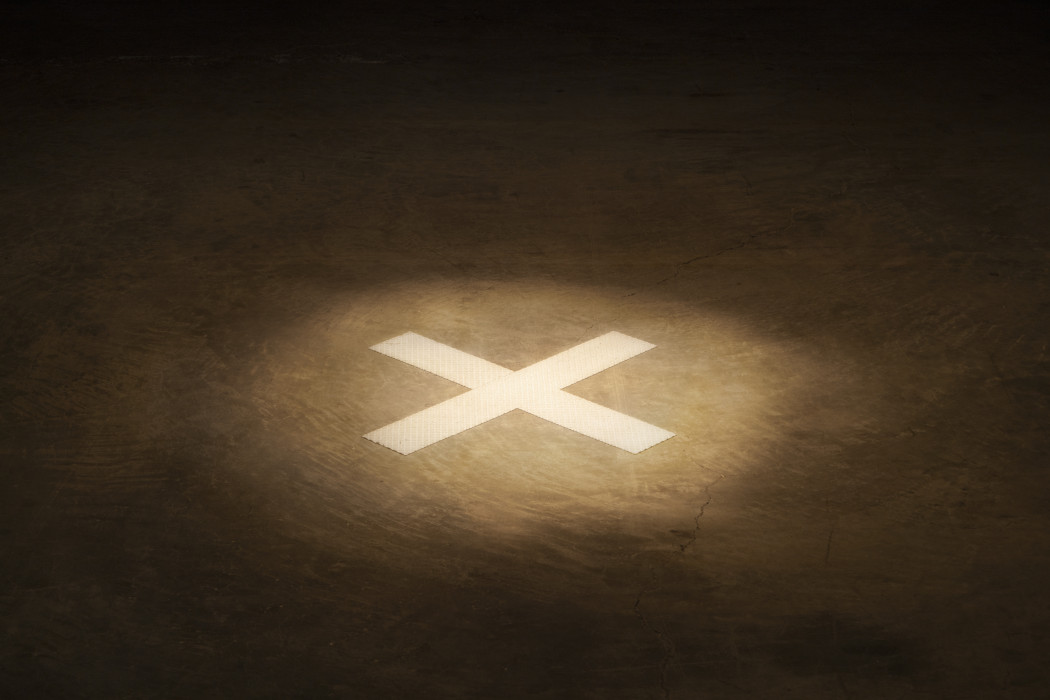 An 'X' made by reflective white tape is spotlit on a dark cement floor.