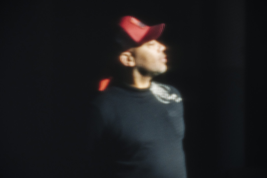 A grainy photo of a person in profile wearing a red baseball cap. The figure is heavily shadowed, the background is black.