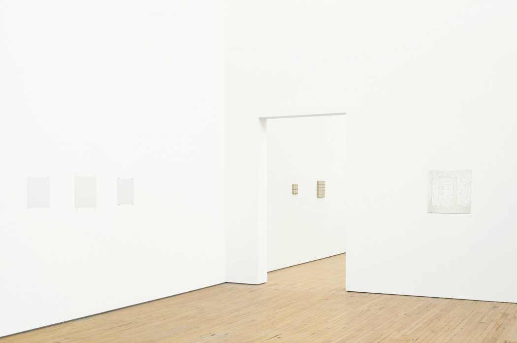 Six small paintings hang on white walls above a wood floor. Three whites paintings are on a wall to the left, one white painting is on a wall to the right, and two tan paintings are visible through a doorway in the center.
