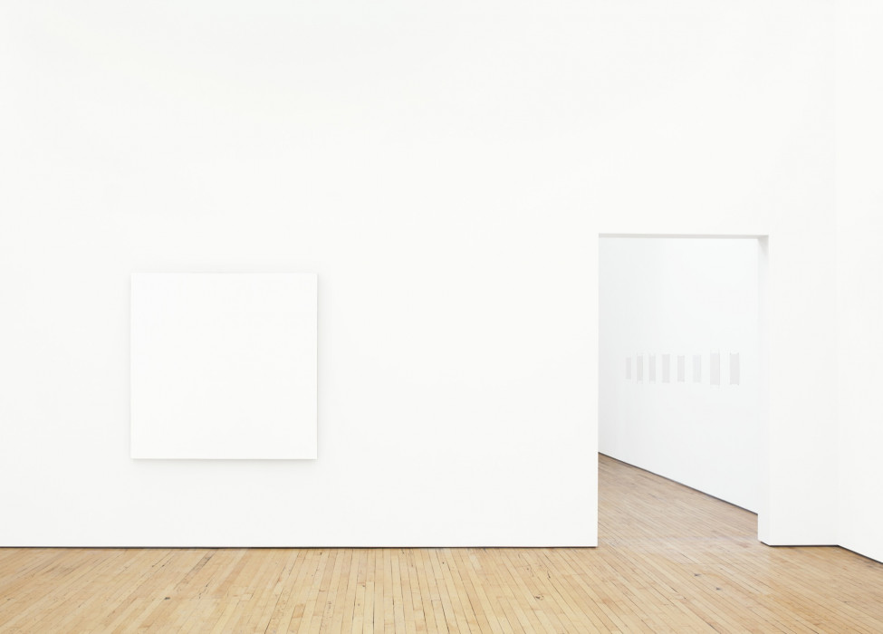 A large, square white painting hangs on a white wall above a wood floor. Eight small, square white paintings are visible through a doorway to the right.