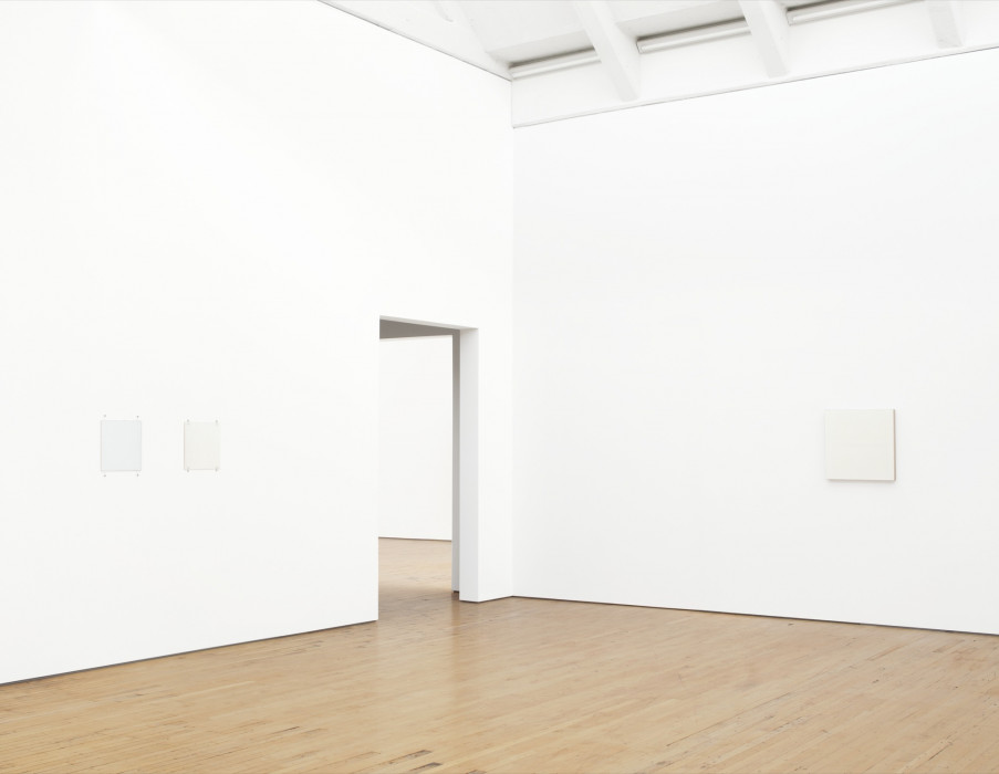 Three small, square white paintings hang on either side of a corner between which is a doorway. The two on the left wall are affixed with fasteners and bolts. The canvas of the larger painting on the right is visible on its right side.