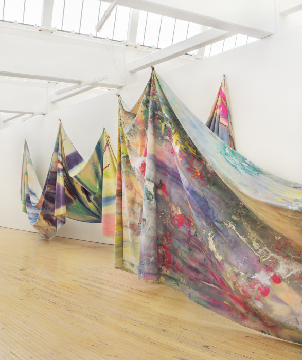 Two massive swathes of multicolored canvas are tied at various points and strung up from the ceiling of an airy, bright, white room. The canvases are painted and splattered with an array of vibrant, pastel, and metallic colors. The artwork floats above the floor.