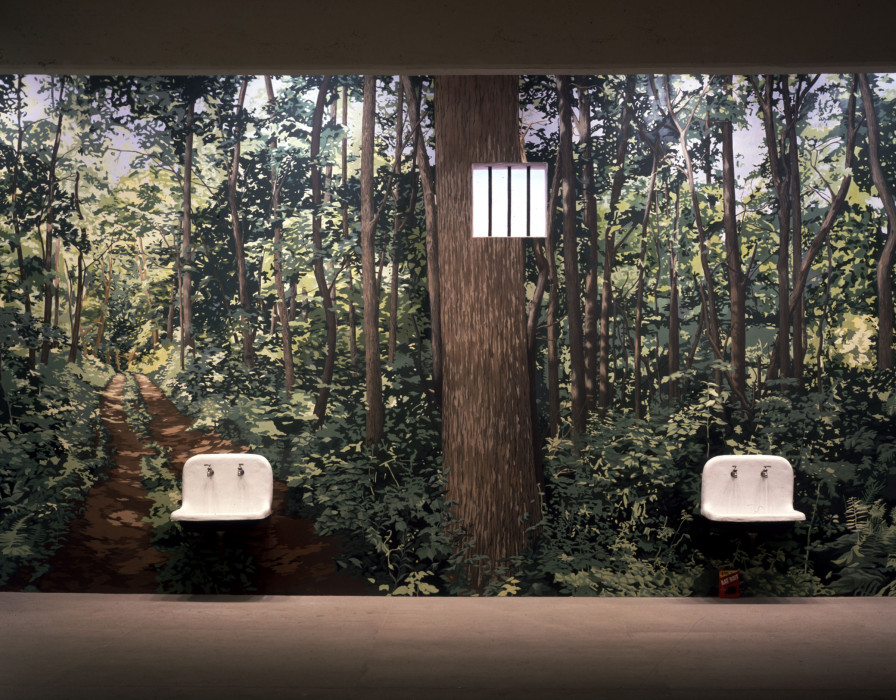An image of a rainforest, covering the whole of a wall in a dark room, with two white sinks installed on the wall near the black floor, and a small white window with vertical grates installed in between the sinks, near the ceiling.