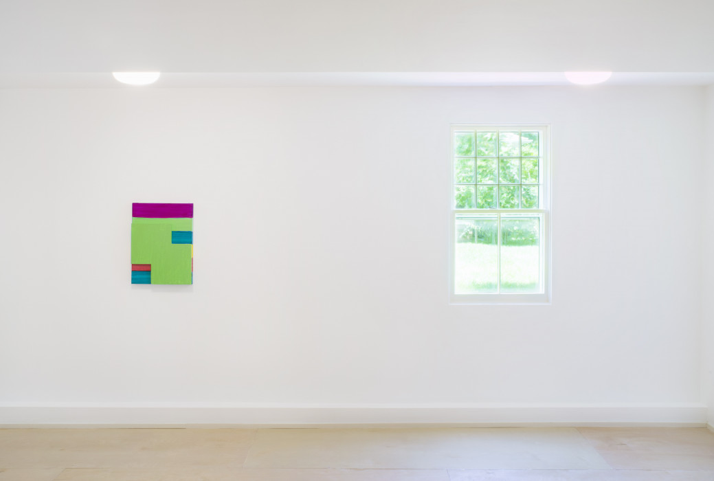 A mutlicolored, portrait-oriented painting hangs on the lefthand side of a white wall and a window is on the righthand side. The painting has horizontal stripes of fuchsia, lime green, teal, yellow, scarlet, and teal sit top to bottom. A lime green shape of two overlapping squares sits on top of the painting.