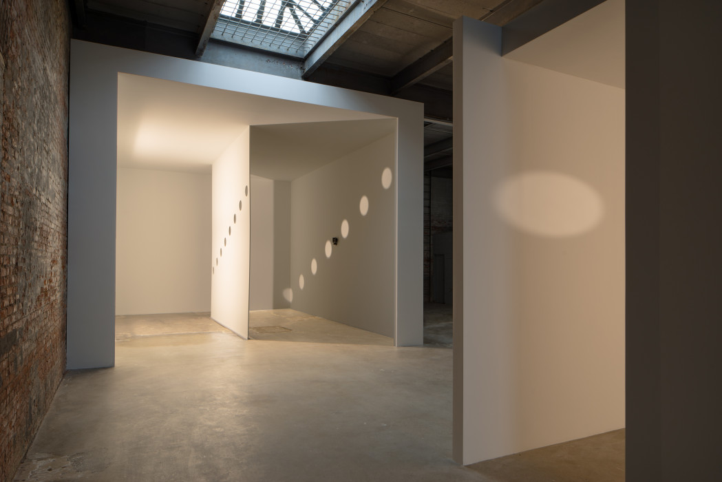 A white room made within an industrial space, with a very thin white wall in the center with eight round holes in it, descending to the left. The light from one side of the thin wall casts shadows of the holes onto the other side of the room.