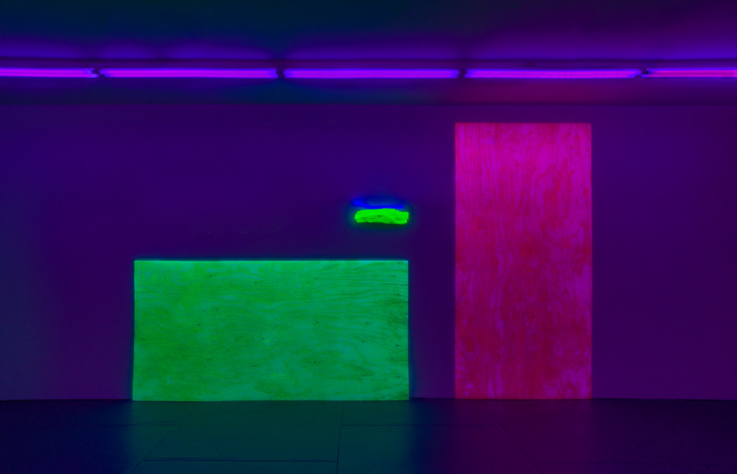 A long horizontal fluorescent green slab of plywood leans against a wall to the left of a tall vertical fluorescent magenta slab of plywood. Projected over the rop right corner of the green slab of plywood is a fluorescent neon green rock form with a thin fluoresent blue worm form sitting on top of it. The room is dark and illuminated by a line of violet fluorescent lights on the ceiling.