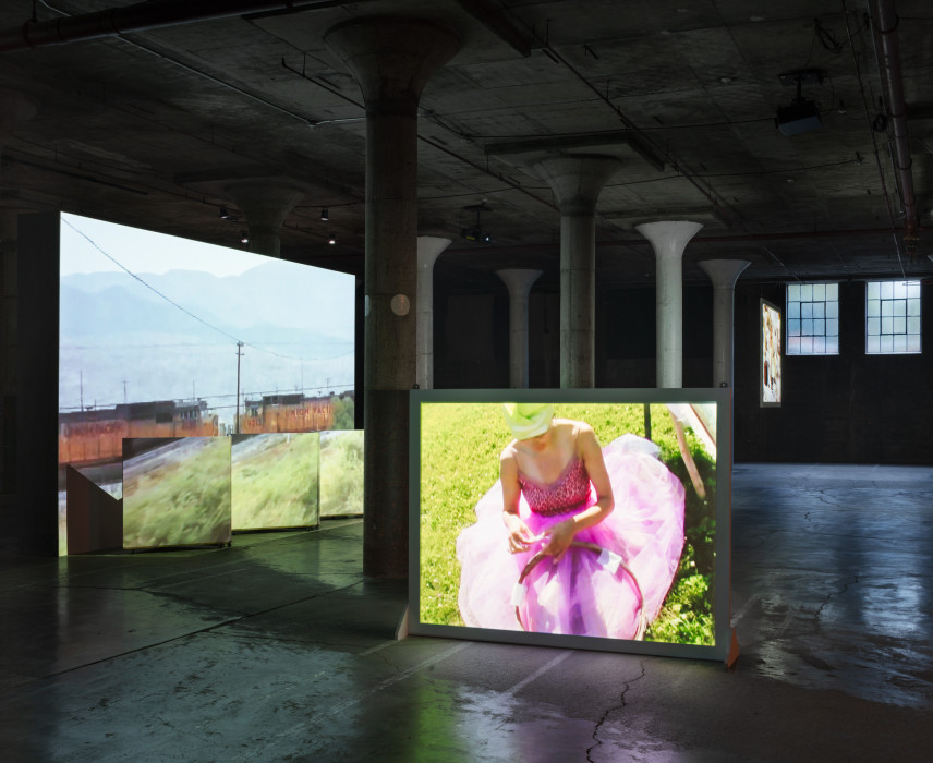 Multiple large screens show projections of videos in a dark space with many columns. Scenes depicted include trains and a person in a pink dress.