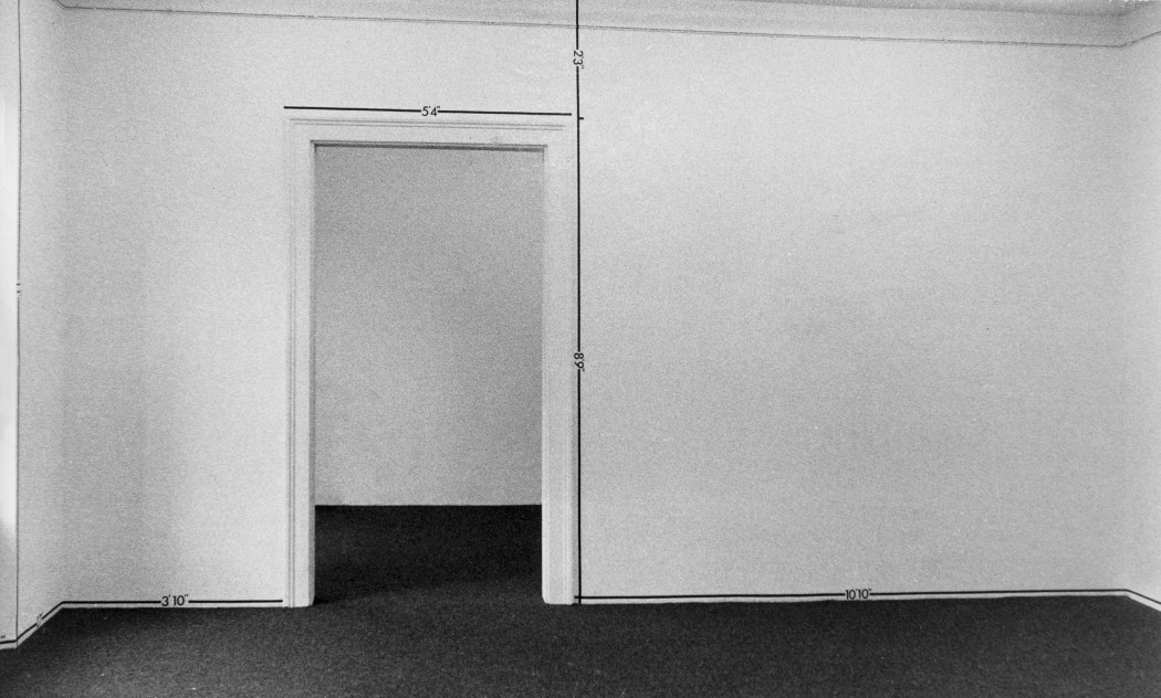 A black-and-white photograph of a white room with a doorway, the room is marked by lines and notes of measurement.