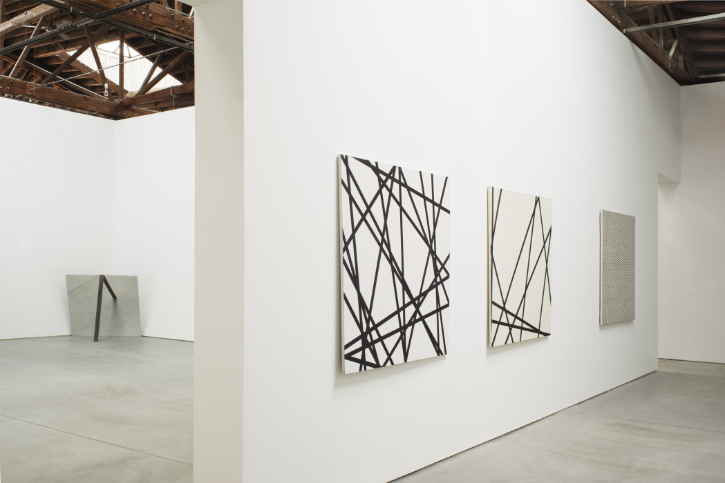 Three square paintings with black-and-white geometric subject matter hang on a white wall. In the background a square mirror sits in a corner and leans forward against a vertical steel bar.