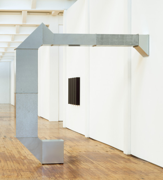 A tall, industrial steel, square tube extends out from a white wall at a right angle and extends towards the ground in another right angle before curving to touch the floor.