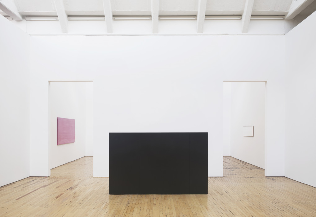 A black rectangular sculptures sits on a wooden floor in between two doorways, with a pink rectangular painting visible through the left one, and a small white rectangular painting visible through the right.