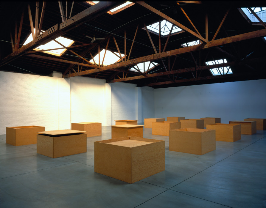 Fifteen plywood boxes arranged in a five rows of three in a large and sunny industrial space, each slightly different.
