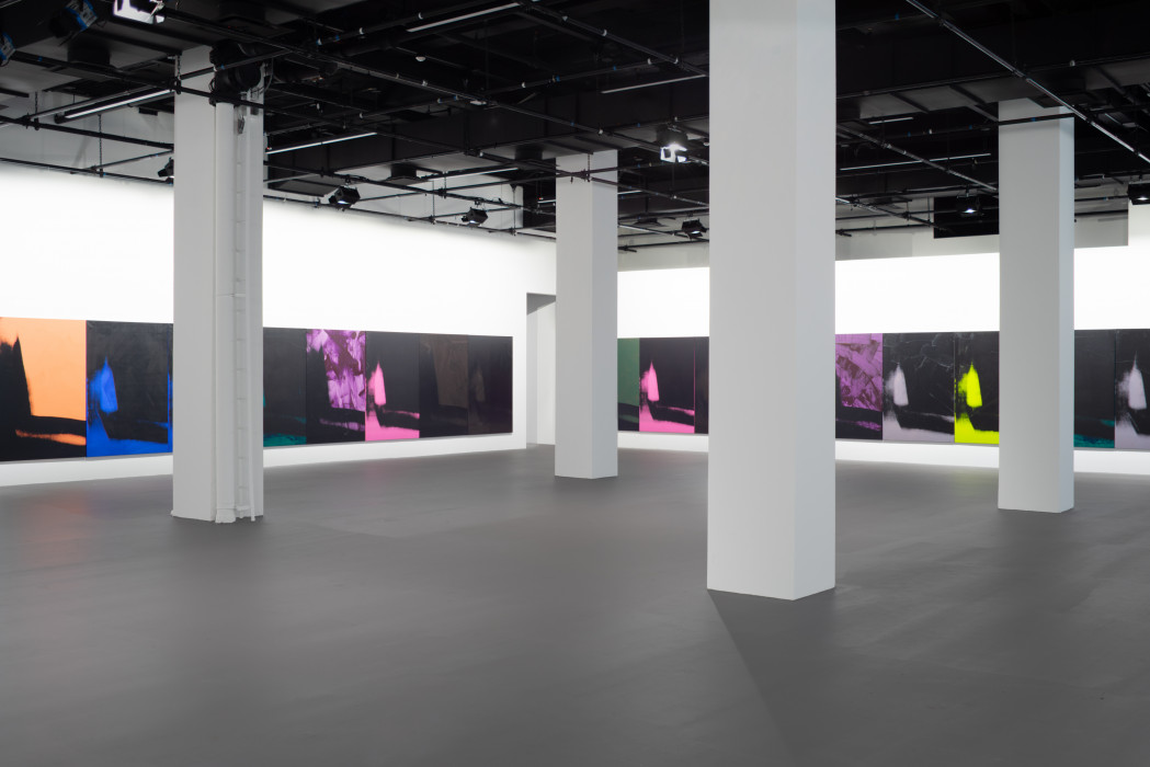 The corner of a gallery with four gray pillars in the center, with both walls lined with many iterations of the same vertically oriented print of an abstracted and vague shadow form that descends on the left and moves across the bottom edge. Each print is in black and one other color, either purple, pink, blue, green, grey, tan, or highlighter yellow, with visible ink strokes and swipes on the purple ones.