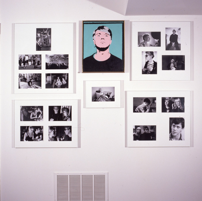 A self portrait silkscreen in blue, pink and black of Andy Warhol surrounded by framed black and white photographs.